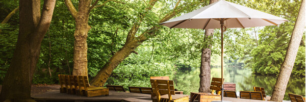 Holzterrasse mit Sitzgelegenheiten und Sonnenschirm am ruhigen See im grünen Wald.